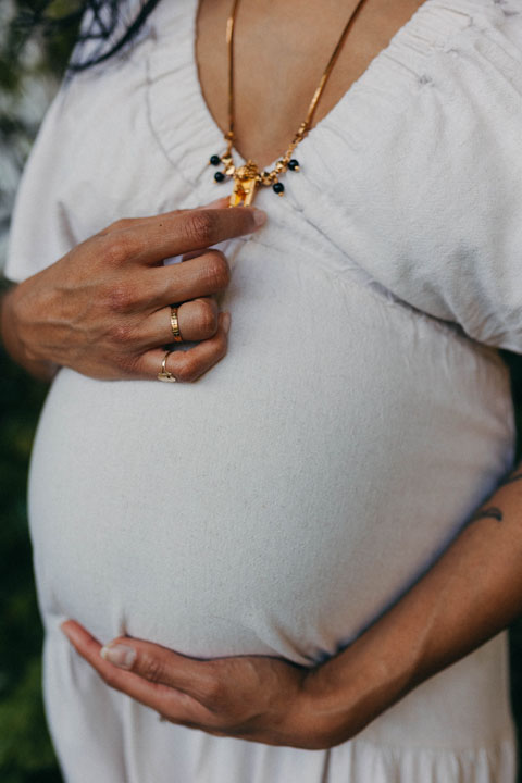 Emotive natural light detail of expectant parents cradling baby bump during jacaranda maternity session
